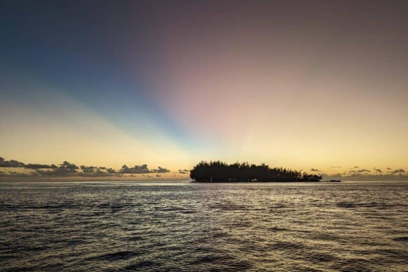 Der Sonnenuntergang über Bora Bora färbt den Himmel orangerot und pastelllau während die darunter liegende Lagune und ein Motu langsam ins Dunkle eintauchen