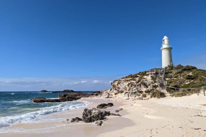 Weißer Leuchtturm auf einem Hügel an einem weißen Sandstrand mit Meeresbrandung auf Rottnest Island in Westaustralien