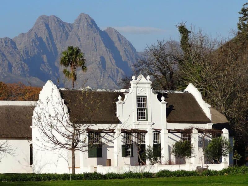 Weißes Kolonialhaus mit braunem Reetdach und kargen Bergen im Hintergrund im Zentrum von Stellenbosch