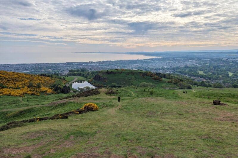 Gelber Stechginster und ein kleiner See auf dem Anstieg zum Arthur's Seat mit Edinburghs Vorstädten und dem Meeresarm Firth of Forth im Hintergrund